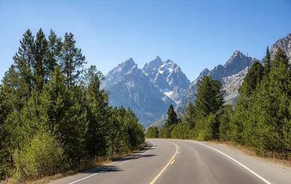 Country road through forest, rugged mountain peaks behind, Grand Teton, Mount Teewinot and Mount St. John, Teton Range mountain range, Grand Teton National Park, Wyoming, USA