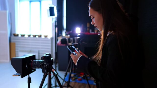 Long-haired Caucasian woman takes video of a camera on tripod on her mobile phone. Footage backstage in the studio.