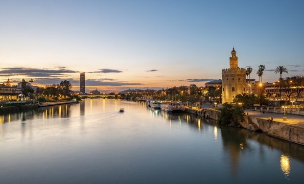 View over the river Rio Guadalquivir with Torre del Oro, promenade and Puente de Triana, in the back Torre Sevilla, sunset, blue hour, Sevilla, Andalusia, Spain