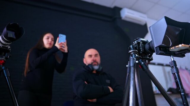 Bald man in black hoodie sits with hands crossed on his chest. Camera on tripod is in front of the cameraman. Low angle view. Photo studio backstage. Lady uses phone in the backdrop.