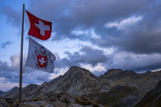 Swiss flag and flag of the SAC with cloudy sky at blue hour and Engadine mountains, St Moritz, Engadine, Grisons, Switzerland