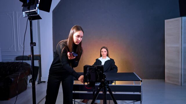 Brunette woman arrange camera on tripod. Cameraman approaches the equipment. Lady site at desk in front of camera. Vlog footage in studio.
