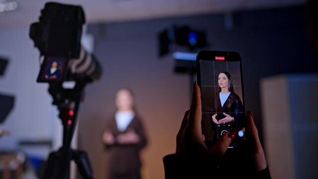 Female hands hold a phone taking video of a beautiful brunette lady. Footage in studio. Blurred backdrop.