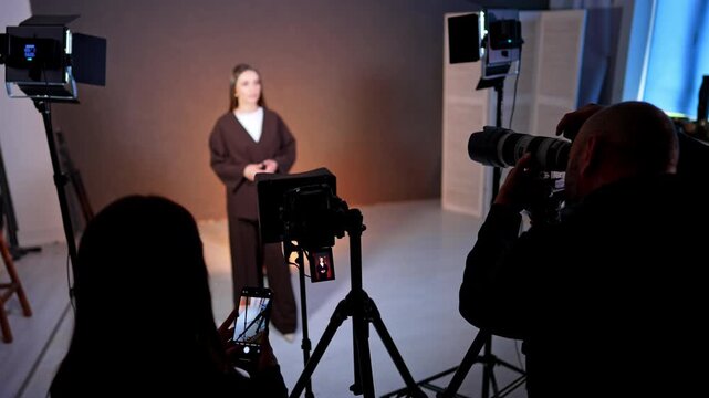 Man and woman are working backstage in photo studio. Woman in brown suit stands in the light of soffits talking to camera.