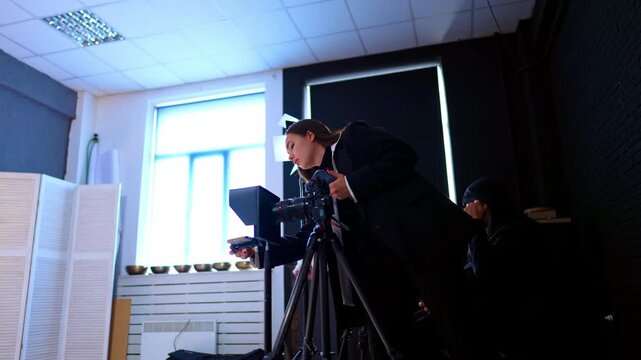Caucasian woman in black suit stands near the camera on tripod. Low angle view at the people working backstage in the studio.
