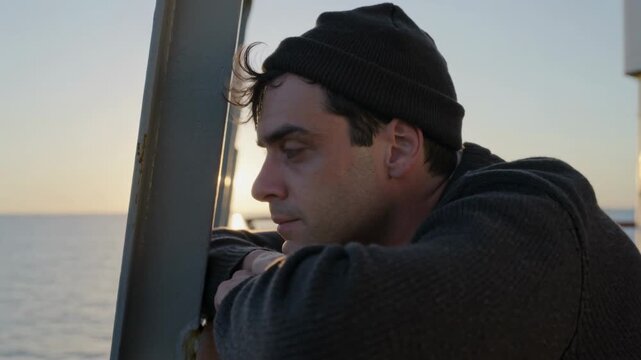 Leaning adult man wearing knit beanie and sweater, lifting head and watching sunset on deck railing