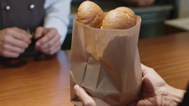 Buyer reaching, server white cuff handing paper bag at wood counter, showing two rolls closing sale
