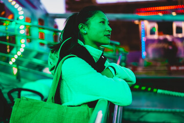 Portrait of young Asian female at carnival, green neon light atmosphere, confident expression, authentic people, inclusion, night amusement park background, creative color, looking away © BullRun
