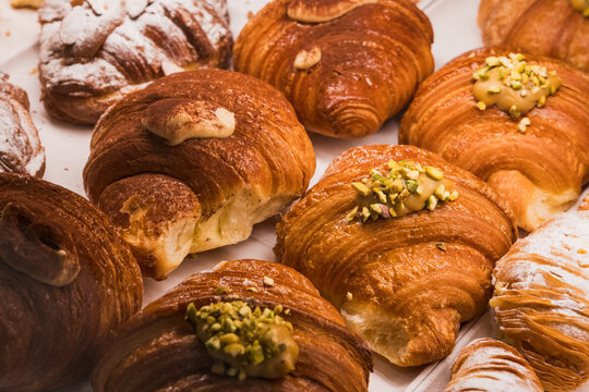Shallow-depth-of-field close-up of a bakery display featuring multiple golden-brown croissants and cornetti topped with crushed green pistachios, cream filling, and powdered sugar. Warm tones, flaky l