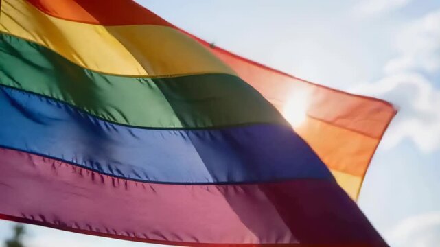 Waving rainbow pride flag letting sun glare through thin fabric under open sky, wind causing waves
