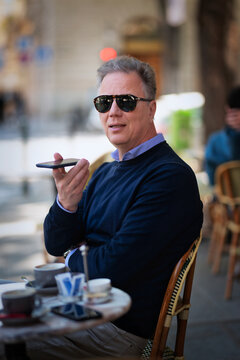 Confident mature man with smartphone sitting at table at sidewalk cafe