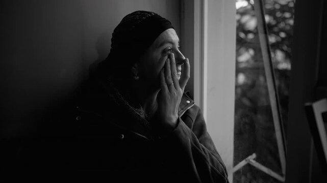 Woman undergoing cancer treatment wipes tears by window, black and white scene capturing grief, emotional pain and vulnerability during a quiet moment at home