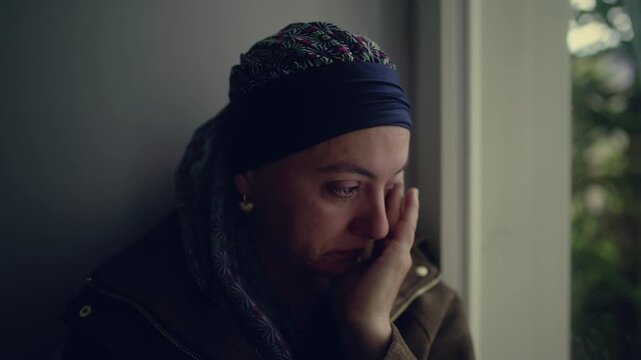 Woman undergoing cancer treatment stands by window wiping tears, emotional moment of deep contemplation, isolation and vulnerability during difficult healing journey at home
