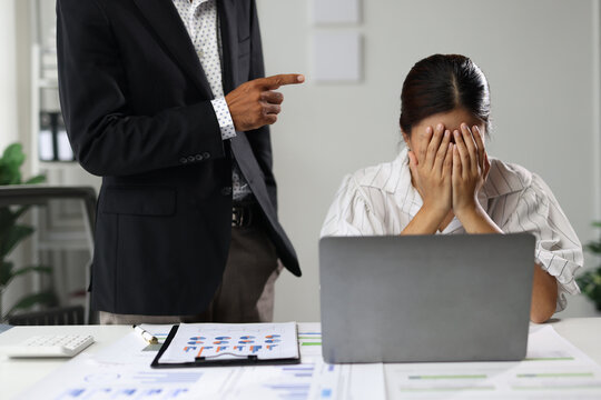 Senior male boss standing over a young Asian female employee, pointing and scolding her, causing her to hold her head in frustration and stress at her office desk during a difficult work situation.