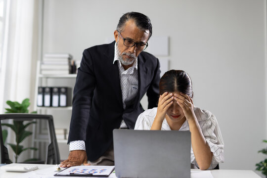 Senior male boss standing over a young Asian female employee, pointing and scolding her, causing her to hold her head in frustration and stress at her office desk during a difficult work situation.