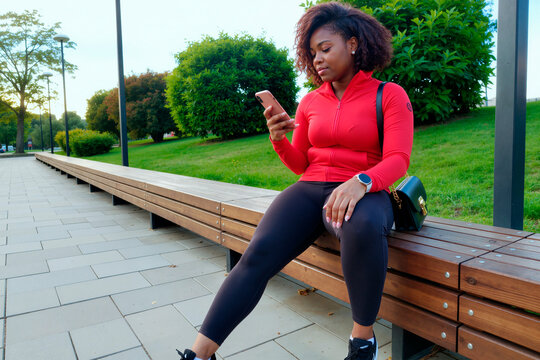 Young African American woman sitting outdoors, checking smartphone after workout and managing mobile communication. Useful for fitness app, wellness marketing, digital lifestyle ads
