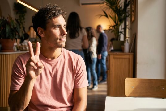 Young man sitting in a cafe, making a peace sign and looking away, with people in background