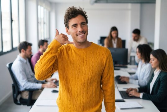 Smiling man in mustard sweater making call me gesture in a modern office with colleagues