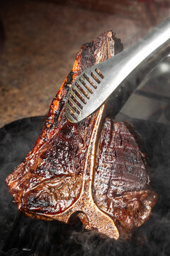Grilled t-bone steak being flipped with tongs on hot pan, juicy seared crust