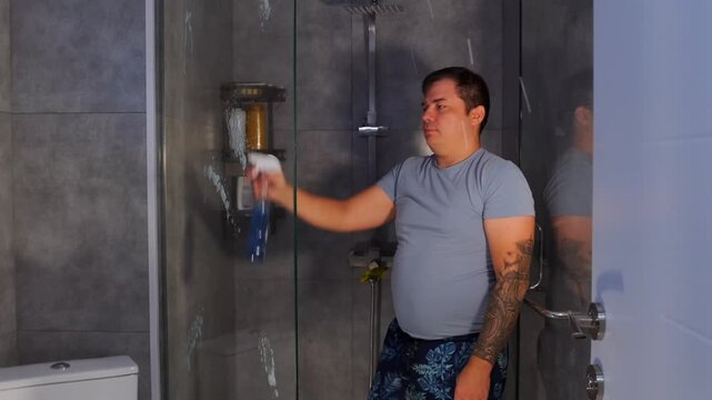 Man spraying cleaning solution onto glass shower wall, removing water stains and soap residue. Domestic bathroom cleaning routine focused on hygiene and maintaining clear glass surfaces
