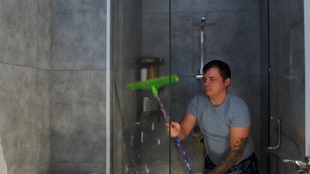 Man using a squeegee to clean glass shower wall, removing water drops and soap residue. Bathroom maintenance routine focused on hygiene, clarity, and spotless surfaces