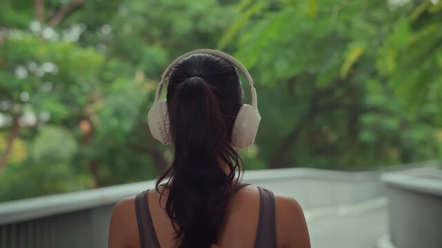 Rear view of a young Asian woman in athletic wear putting on wireless headphones in a lush green outdoor park. This scene captures mindfulness, mental wellness, and a healthy lifestyle through music