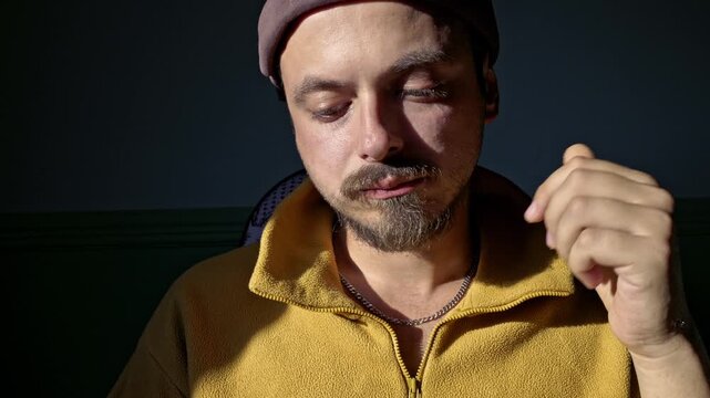 Close-up of a bearded man in a yellow fleece jacket and a beanie, focused on eating a small snack in a dimly lit indoor setting.