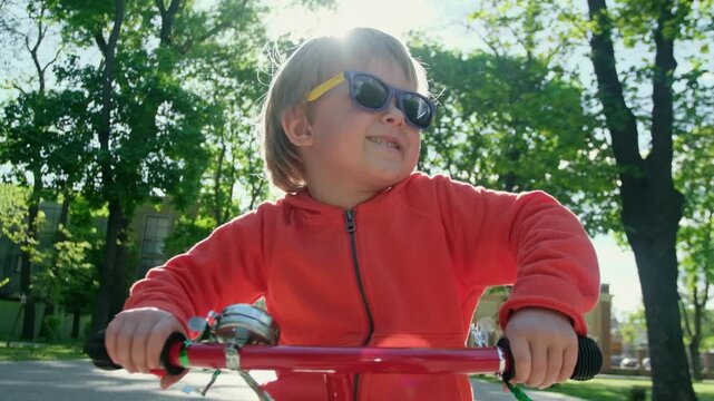 A happy child in sunglasses holds the handlebars of his bicycle and smiles happily while looking at the camera against the backdrop of the setting sun in the park.