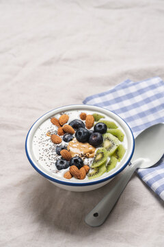 High protein breakfast with yogurt, chia seeds, almonds, blueberries, kiwi and peanut butter in light bowl on linen tablecloth