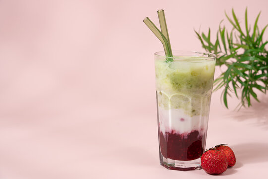 Strawberry matcha latte in clear glass on pastel light pink background with copy space, layered iced drink, minimal aesthetic
