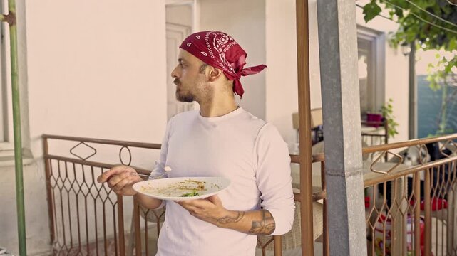 A young man with a red bandana on his head is seen enjoying a simple and delicious meal on his home balcony. He savors every bite while taking a casual break outdoors.