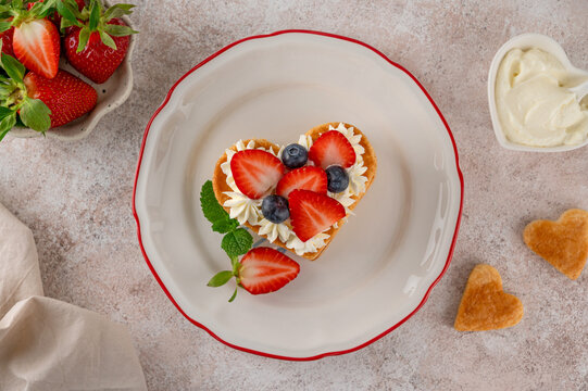 Valentines day dessert. Heart shaped cake millefeuille with vanilla cream and fresh berries on a plate on a light concrete background. Romantic food concept. Top view.