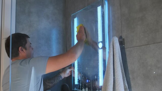 Middle aged white man wipes bathroom mirror with cloth, removing stains and fingerprints. Household cleaning routine, hygiene care, reflective surface polishing and everyday domestic chores at home