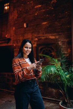 Smiling hispanic woman using smartphone outdoors, dark haired female browsing social media application, young lady holding mobile phone in city, mixed race model using wireless 5G data connection.