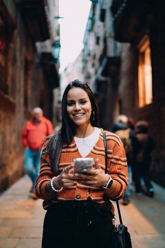 Happy multiracial female user with smartphone in hands outdoors, young lady texting on mobile phone in narrow city alley, hispanic woman using wireless data connection, authentic representation.
