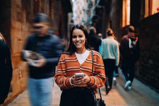 Smiling hispanic woman using smartphone on busy city street, young female with mobile device standing in crowd, mixed race model with wireless connection, lifestyle of modern digital nomad.