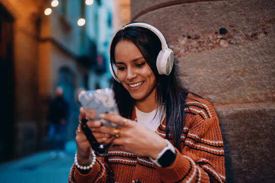 Happy multiracial girl texting on mobile phone wearing headphones, young hispanic female using smartphone audio app in urban setting, lady checking social media while listening to podcast.