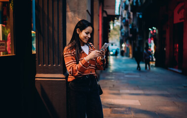 Fototapeta premium Smiling Hispanic woman leaning against wall using smartphone, night city street alley, browsing social media, mobile app usage, digital nomad lifestyle, authentic portrait, communication.