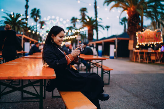 Cheerful mixed race woman browsing on mobile phone while sitting outdoors, happy hispanic girl using smartphone application at evening fairground, authentic digital nomad life, copy space.
