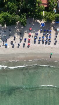 Imagem a&eacute;rea de praia com guarda-s&oacute;is, banhistas e ondas quebrando na areia, representando lazer e turismo em cen&aacute;rio tropical brasileiro