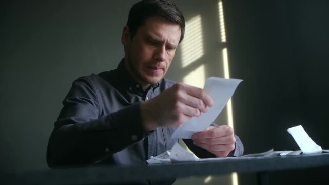 Serious man sitting at table checking his household expenses and receipts, looking worried about his financial situation, calculating costs and payments during an economic crisis. 