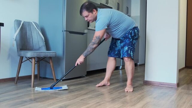 Man mops wooden floor in modern kitchen, removing dirt and dust with wet mop. Household cleaning routine, hygiene, domestic chores, maintenance and everyday home lifestyle activity
