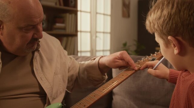 Zoom out shot of child learning to play electric guitar at home and pointing at headstock with grandfather or music teacher helping, scene lit with warm light Great for beginner guidance