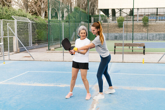 Young female coach guiding an older woman on playing pickleball, showing paddle grip and ball placement on a blue court