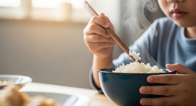 Child enjoying rice with chopsticks in cozy kitchen setting, steamy bowl of rice held carefully. Childlike innocence and daily routine reflected in meal preparation.