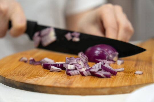 Chef's hands slicing a red onion with a black knife on a rustic wooden cutting board, dicing fresh purple onion for meal prep in a home or restaurant kitchen close-up