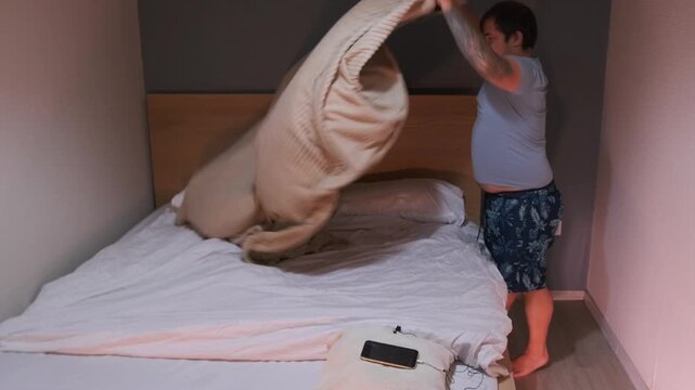 Man spreading bedspread over bed in bedroom, making bed and arranging bedding neatly. Daily household routine, home organization and cleanliness in a modern apartment interior