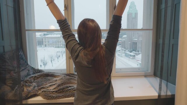 A person enjoying a peaceful morning by the window, stretching and taking in the snowy cityscape outside. The cozy interior setting complements the winter atmosphere, creating a sense of tranquility a