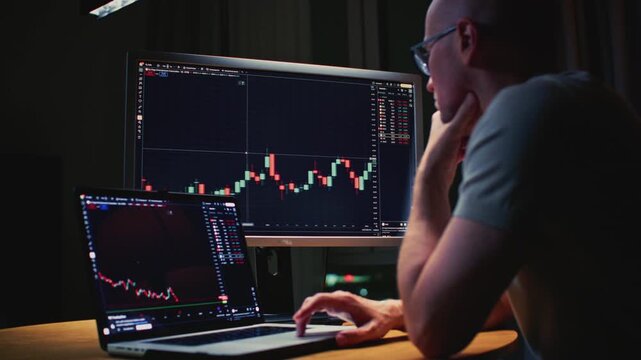 Man trader monitors market charts on laptop, screen and smartphone at home. Analyzing real-time trading data, considering investment strategies. Professional investor engaged in day trading, Evening