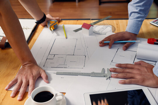 Close up of architect team hands reviewing house blueprint with vernier caliper and miniature model on wooden desk in office.
